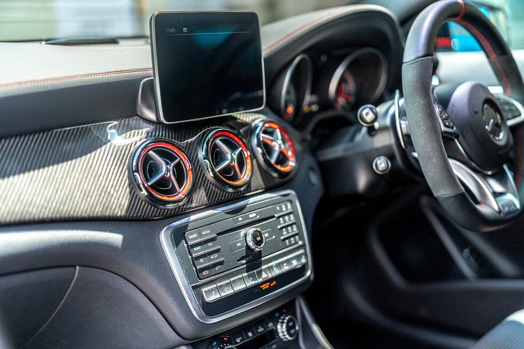 Close-up of a luxury car interior featuring a carbon fiber trim panel, circular air vents with metallic rings, a central infotainment screen, and a multi-function steering wheel.