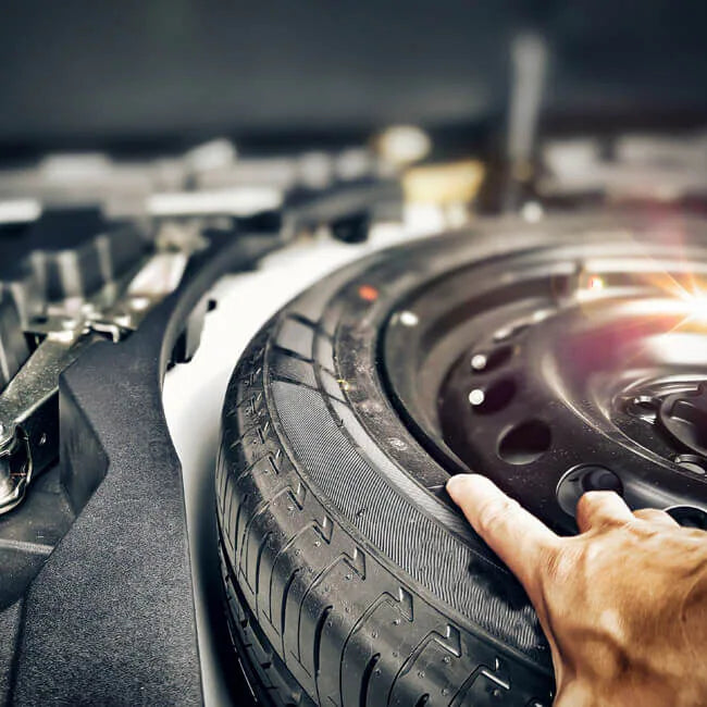 A close-up shot of a spare tire inside a car trunk, with a person's hand resting on the tire sidewall.