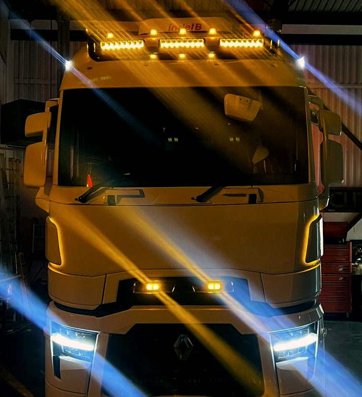Front view of a large truck cab with amber roof-mounted LED light bars and bright white headlights, photographed in a garage with dramatic lens flare effects.