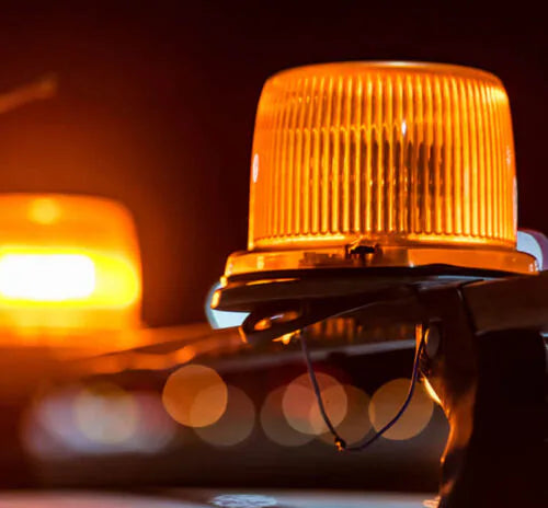 A close-up of a flashing amber emergency vehicle light mounted on a dark vehicle, with a blurred second light in the background.