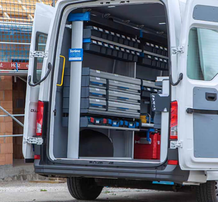 A white work van with its rear doors open, showing a mounted tool organizer system inside. The organizer contains gray and blue storage bins and trays, with a visible Sartimo brand label on a vertical support.